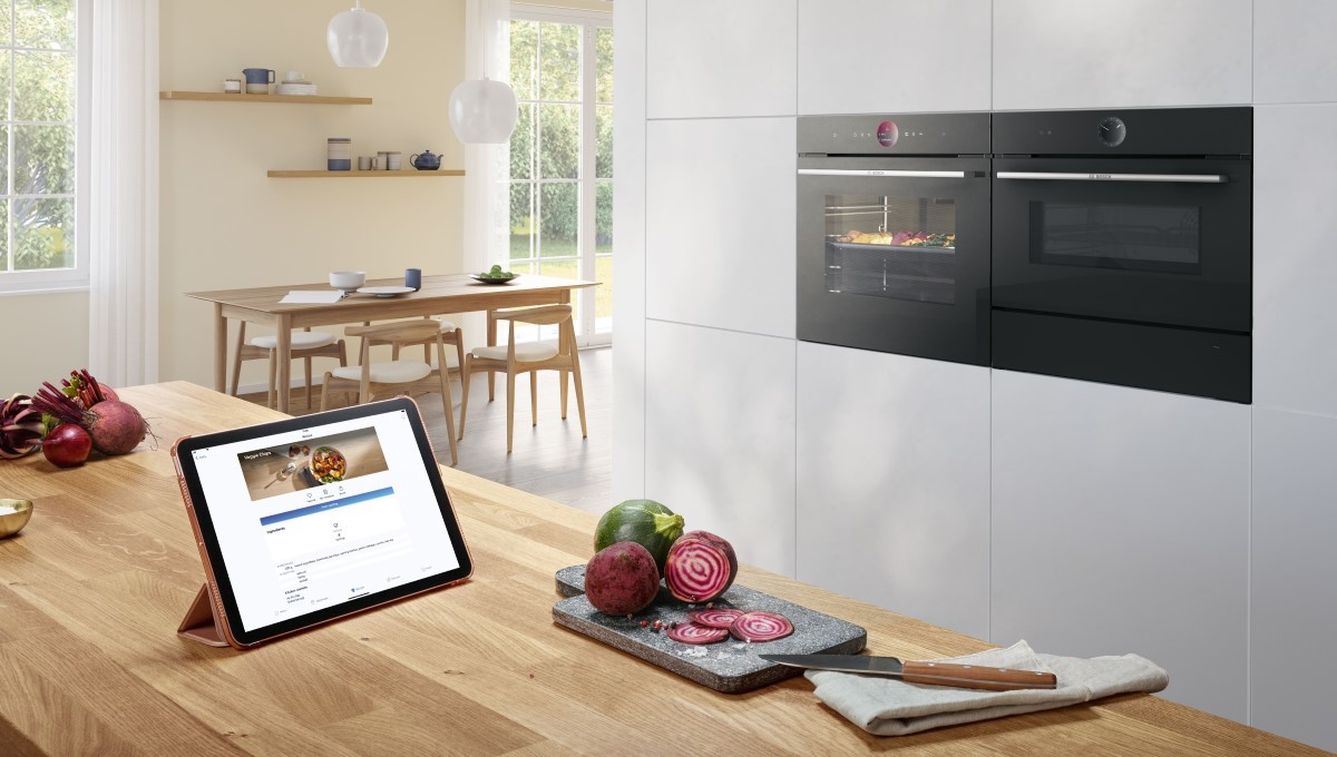 W wysokiej zabudowie piekarnik obok mikrofali. Na przeciw nich stoi stół, na którym leży tablet z aplikacją Home Connect.