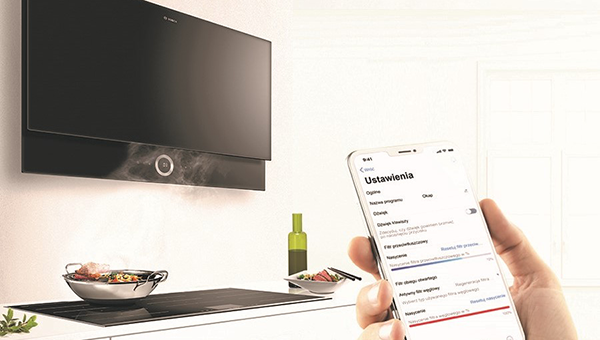 Na pierwszym planie ręka ze smartfonem i wyświetlaną aplikacją Home Connect. W tle okap, który wisi nad płytą indukcyjną.